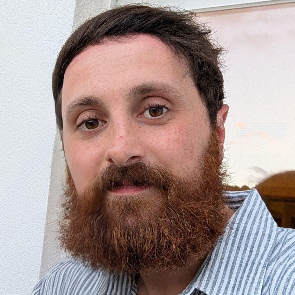 A close-up of a man in a white and grey striped shirt. He has short brown hair and a bushy beard