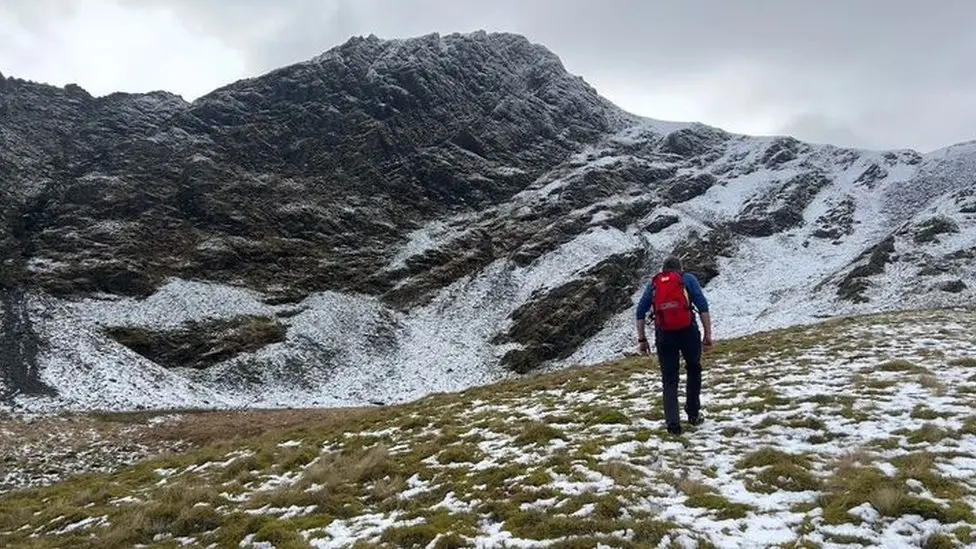 Mountain Rescue team member, wearing a blue top and red rucksack, walks up to Blencathra on the day of the accident. There is frost on the ground and a rocky peak is close by.