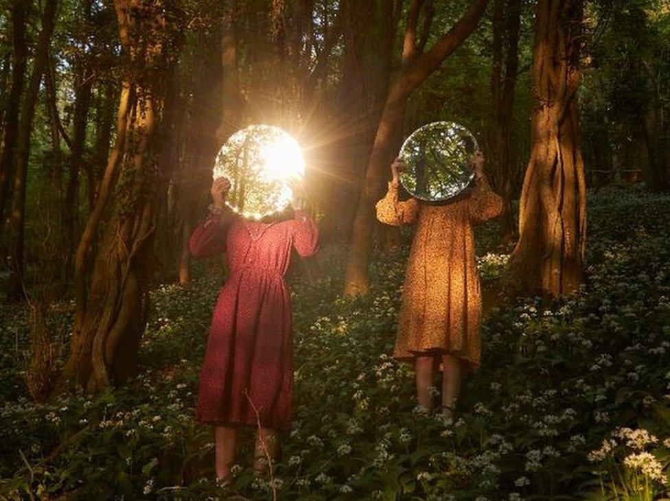 A photo of two women wearing colourful dresses, holding up mirrors which reflect light from their face in the woods.