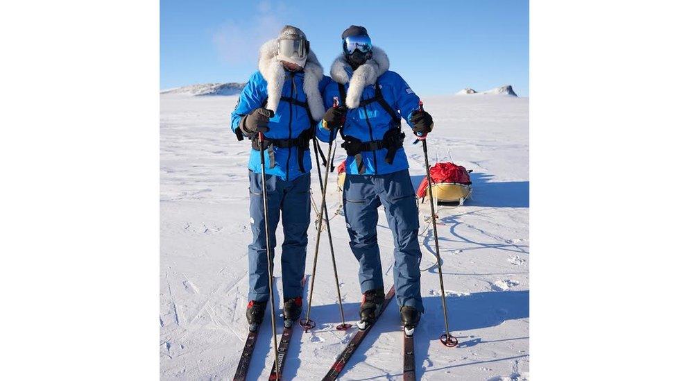 justin and jamie standing together in the antactic wearing skis with their luggage behind them