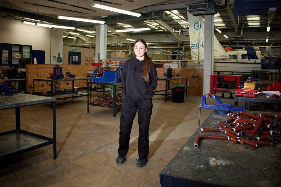 Chloe Green standing in one of the college's workshops. There is a white sailing boat in the background