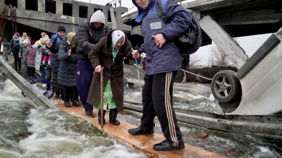 A line of people, including children being carried and an elderly woman with a walking stick, gingerly cross the river amid the wreckage of a broken bridge, walking on planks