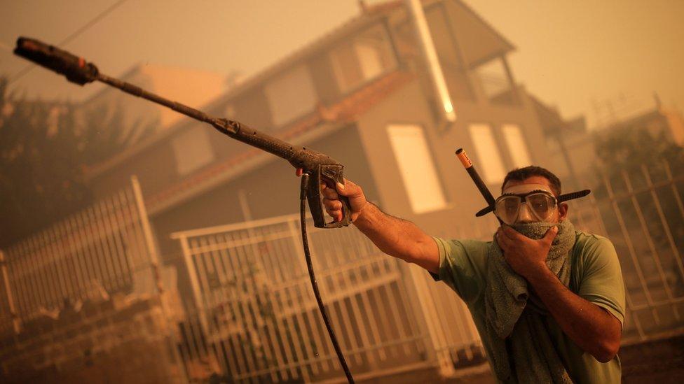 A Greek resident wearing a scuba-diving mask sprays water at the entrance of a house during a wildfire