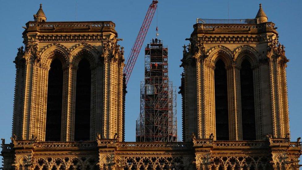 A crane seen lowering a new rooster weathervane into place on the spire of Notre-Dame. The cathedral's two towers are seen either side of the foreground.