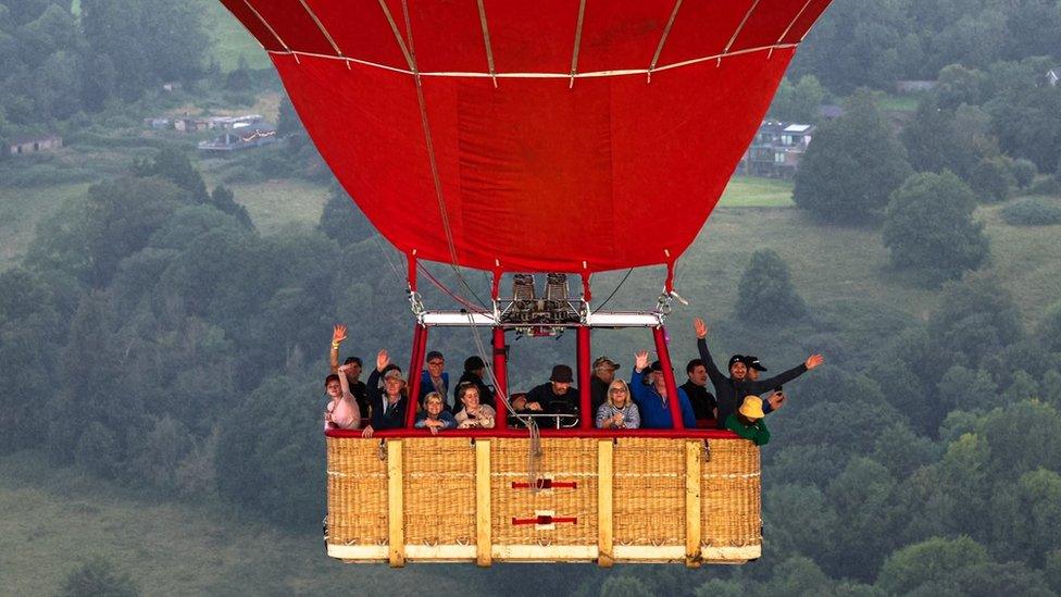 Around 18 passengers waving from the large rectangular basket of red balloon