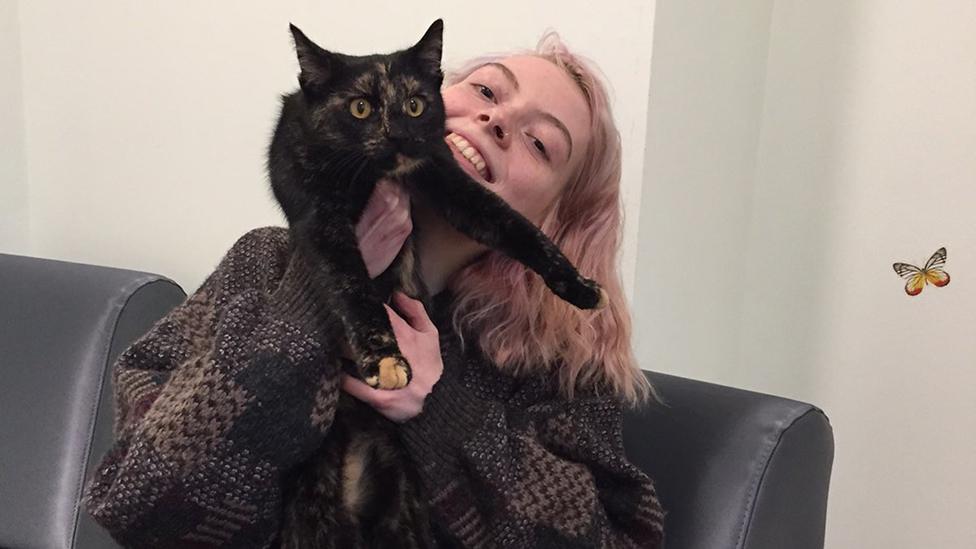 Hat is young, white, and wearing a large, dark chunky cardigan. Hat's got long blonde hair and is smiling, but their face is obscured because Hat's holding up a mostly black, tortoiseshell cat, which Hat is cuddling. The cat looks wide-eyed and a little bit nervous, but it's a happy scene.