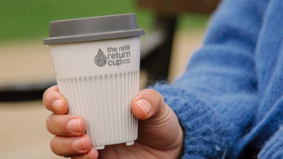 A white cup with a grey lid that is reusable and being held by a woman with a blue jumper on