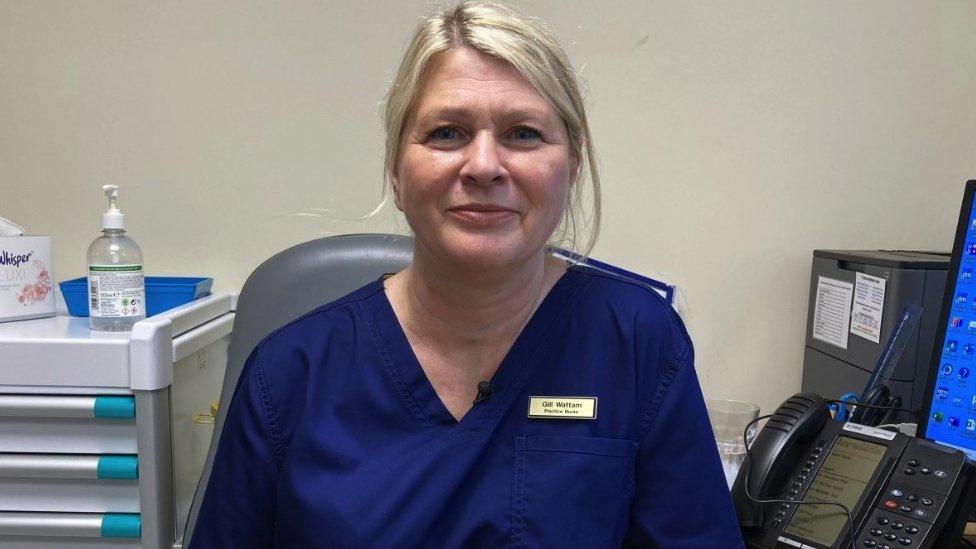 Gill Wattam. She is sat at a desk and a telephone and computer can be seen next to her. She is wearing a navy blue nurse's uniform and a yellow name badge with her name and 'Practice nurse' written below it in black text.