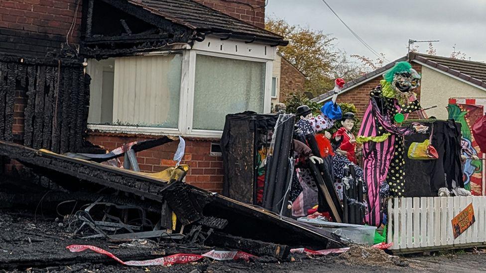 The burnt out remains of the Halloween display at Stan Yannetta's house in South Shields. To the left of the image is a burnt down, black fence. A bay window is also damaged, with the roof black and one side missing. Part of the display is in the garden next to the window, behind a white fence. There are statues of monsters and other Halloween props.