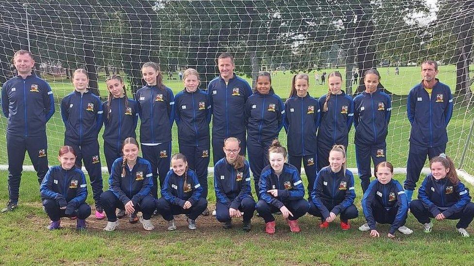 Members of AFC Rumney under-14s girls football team wearing blue tracksuits with the club's crest emblazoned on the front, standing in field in front of football posts