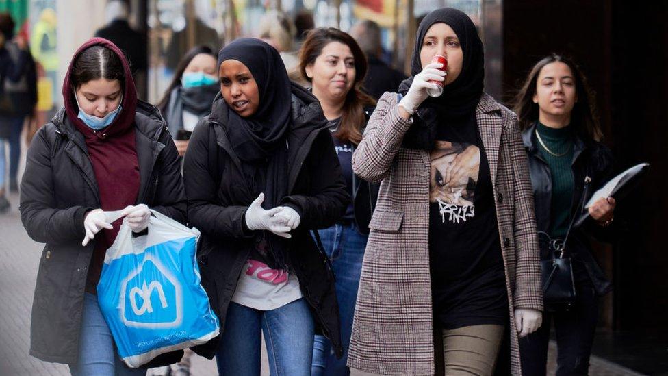 A group of young women with head-scarves walking on the street in The Netherlands. Some wear face masks, some wear gloves, and some wear no protection at all.