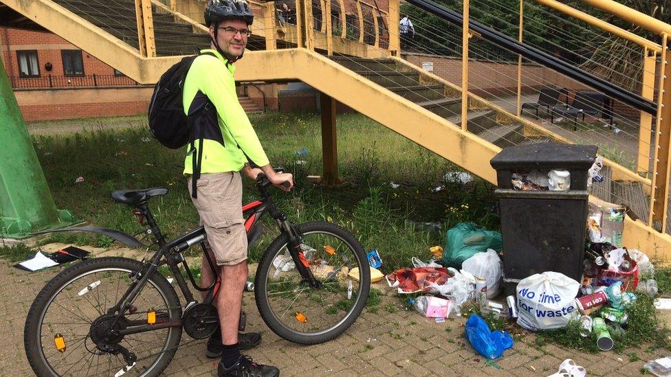 A cyclist stands by his bike, surrounded by litter and an overflowing rubbish bin in Northampton.