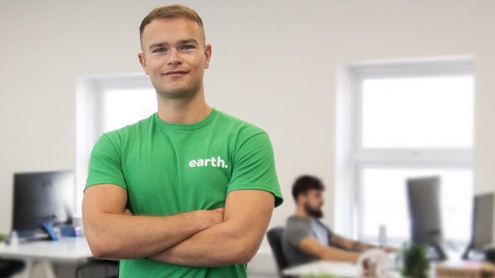 Luke Chapman folds his arms while wearing a green shirt in his office with a worker in the background