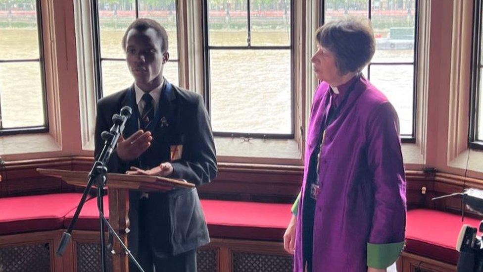 A Bishop in a purple robe and a young black school child speaking into a microphone in an ornate room