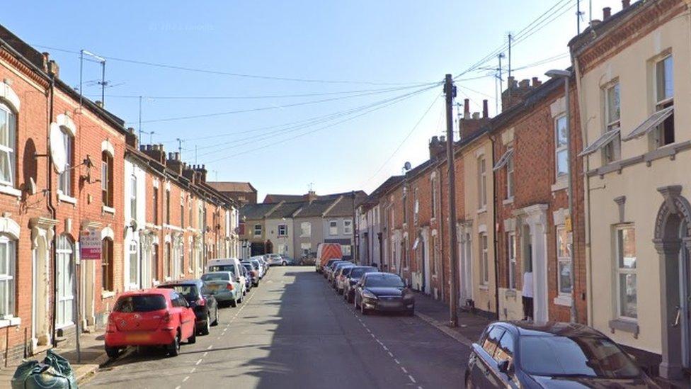 Terraces of early c20th houses with cars parked on either side of the street