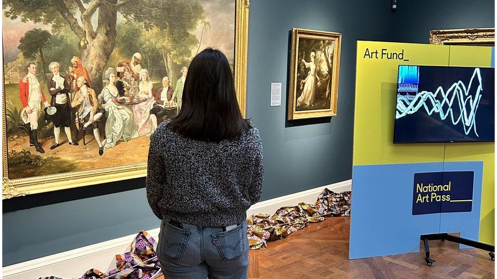 Back of a woman who is standing looking at a painting on a wall at a gallery