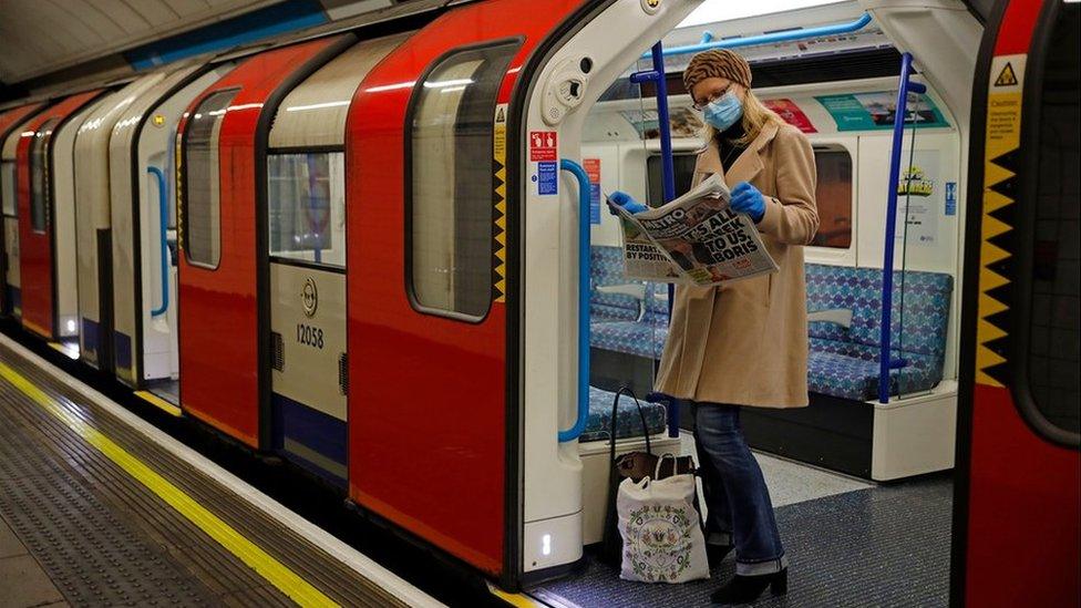 A woman wearing PPE (personal protective equipment), including a face mask as a precautionary measure against COVID-19, reads a newspaper as she stands aboard a London Underground Tube train, i