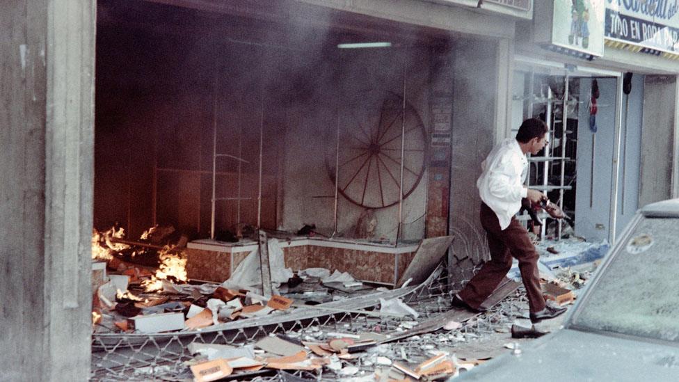 A man with a pair of shoes in his left hand runs out of a destroyed shoe store on February 27, 1989