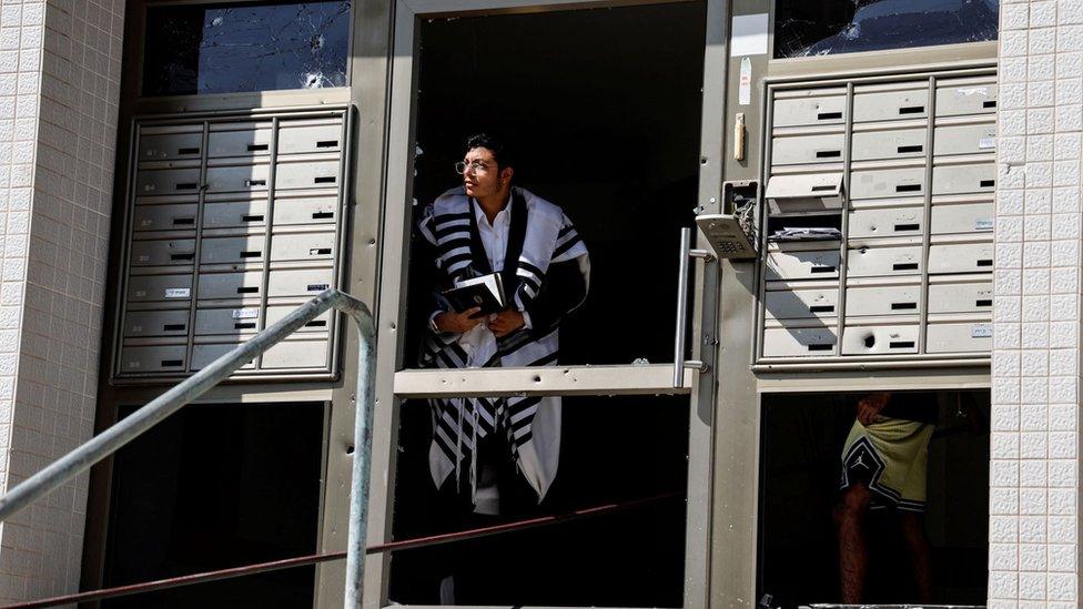 A man wearing a Jewish prayer shawl looks out of the damaged entranceway to a building, as rockets are launched from the Gaza Strip, in Ashkelon, southern Israel October 7, 2023.