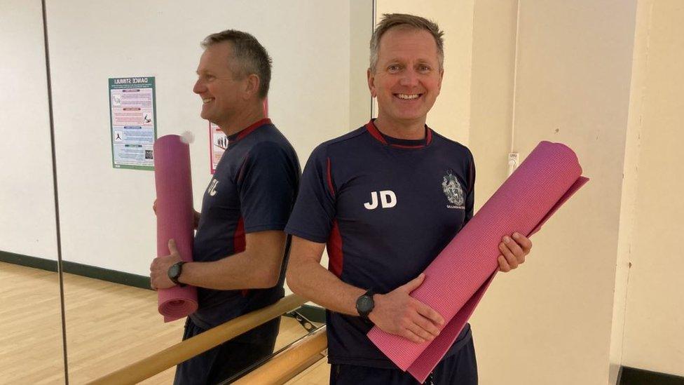 Man stands against studio mirror holding a pink yoga mat