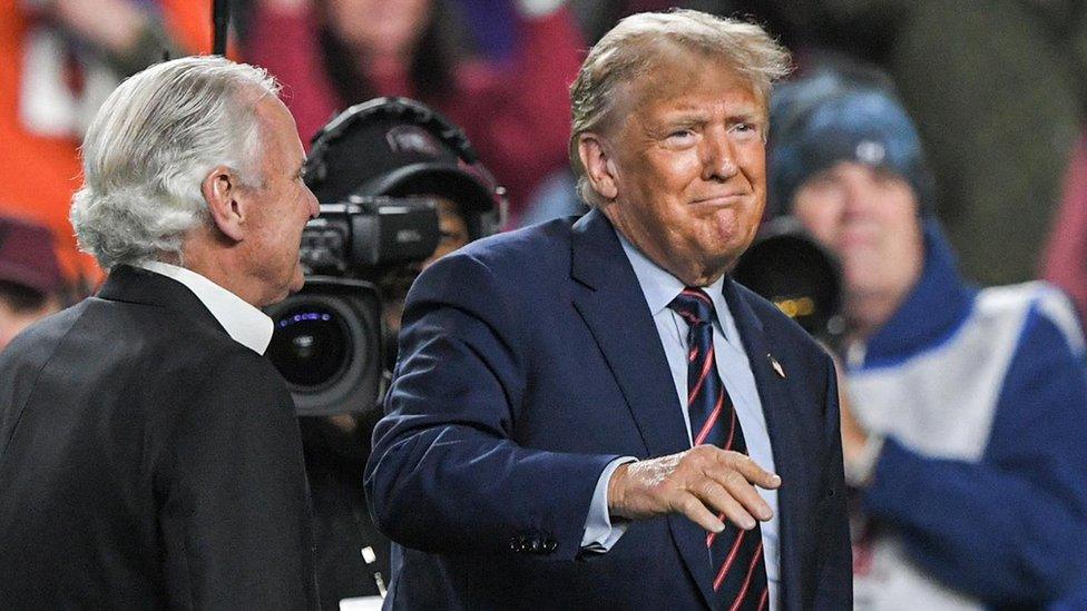 Trump in navy blue suit, hand raised, in front of tv cameras