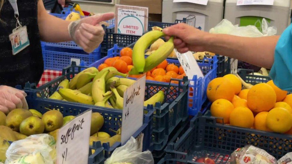 a hand picks two bananas from a table with boxes of bananas, pears and oranges on it