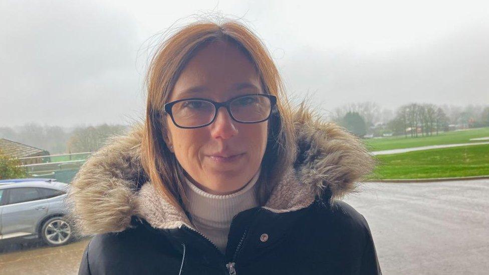 Woman with long fair hair and glasses wearing an anorak with a furry hood stands in a park