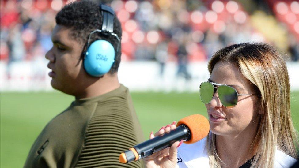 Katie and Harvey at an event. Harvey is wearing ear defenders while Katie talks into a microphone