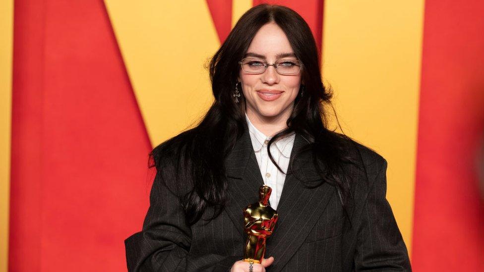 Billie Eilish, wearing a black suit and white shirt, holding her Oscar award at the 2024 Vanity Fair Oscar After Party