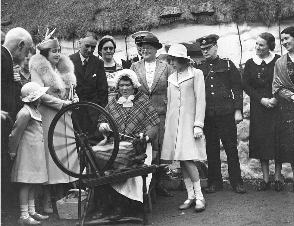 Queen Elizabeth, Queen Consort to King George VI (left) with Princesses Elizabeth (right) and Margaret Rose, watching a spinner at work in a Scottish Highland village.