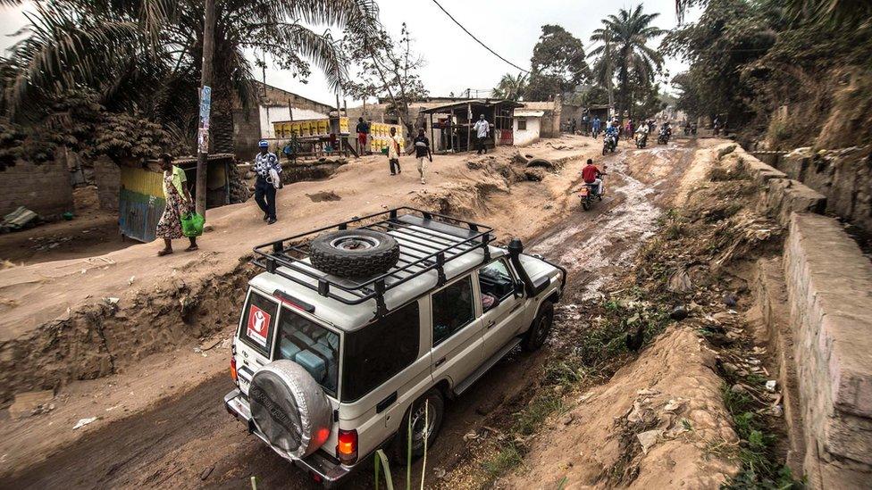 4x4 vehicle drives through dirt track with steep banks either side where pedestrians are walking