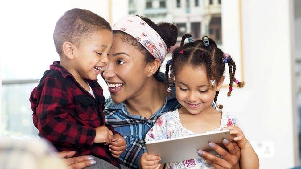 A woman plays with her two children at home with her partner in the background