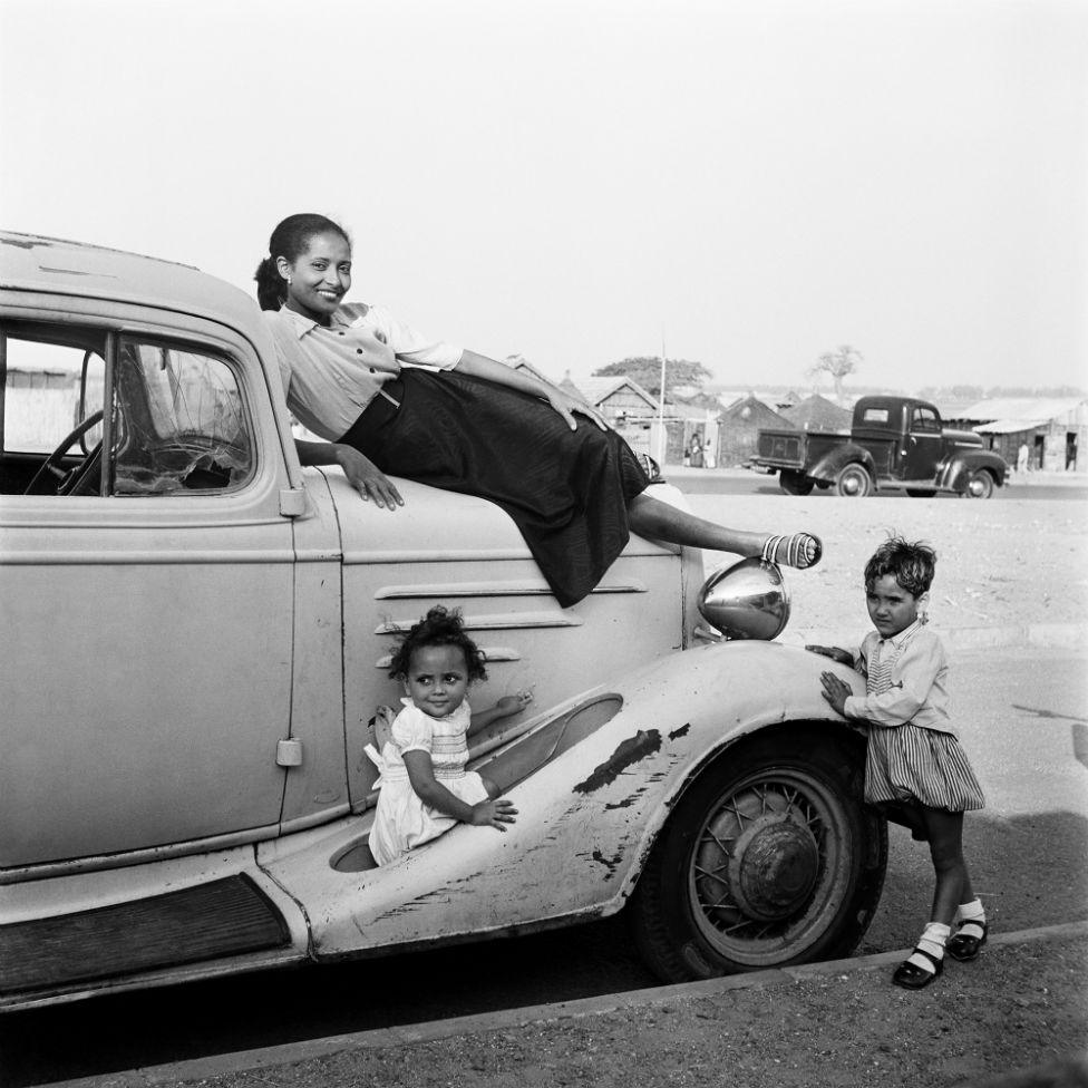 A woman, Madame Gomez, poses on top of a car with two young children standing by the vehicle