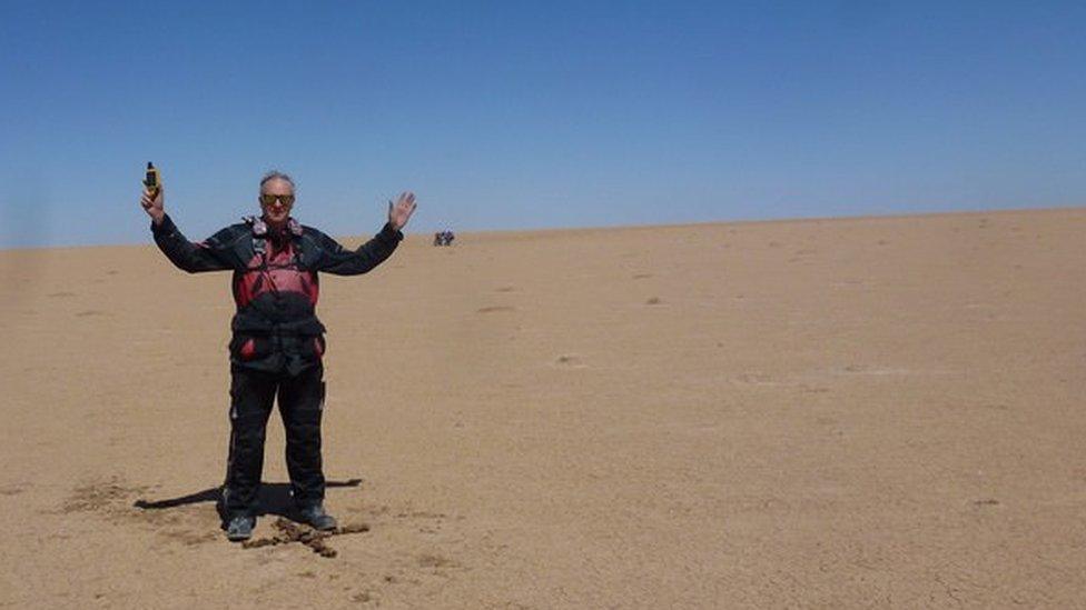 A man stands in the middle of a barren desert, above an 'X' carved into the sand