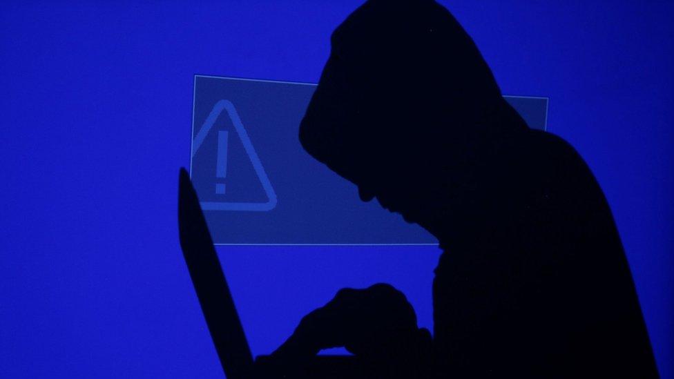 A hooded man holds a laptop computer as blue screen with an exclamation mark is projected behind him