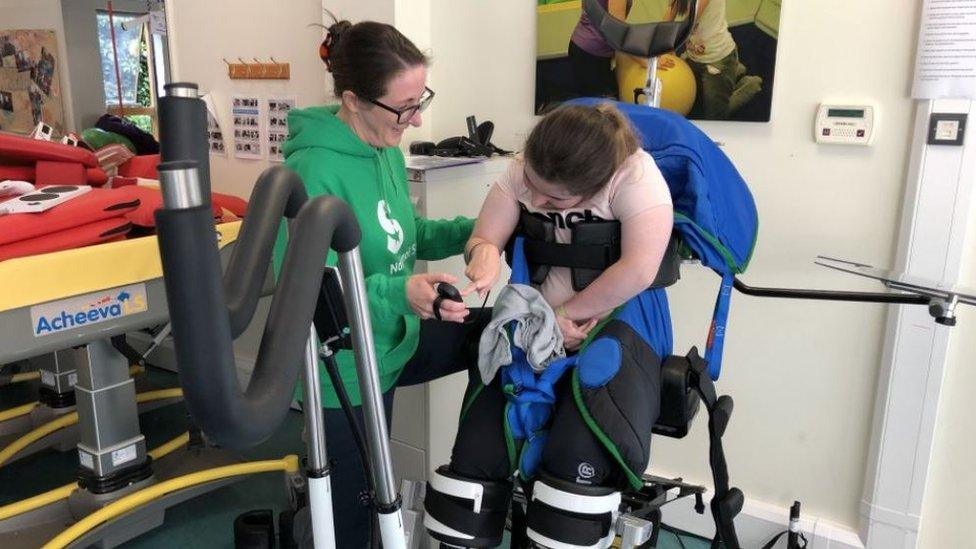 A person in a piece of equipment described as a cross trainer for those who can't walk. Another person is standing at the side helping them.