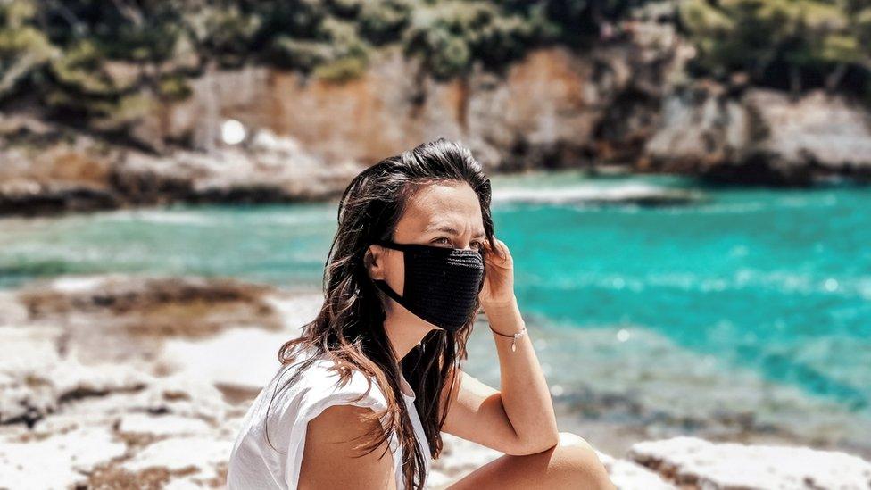 A young woman wearing a face covering, sitting on some rocks in front of blue sea water sea in Croatia