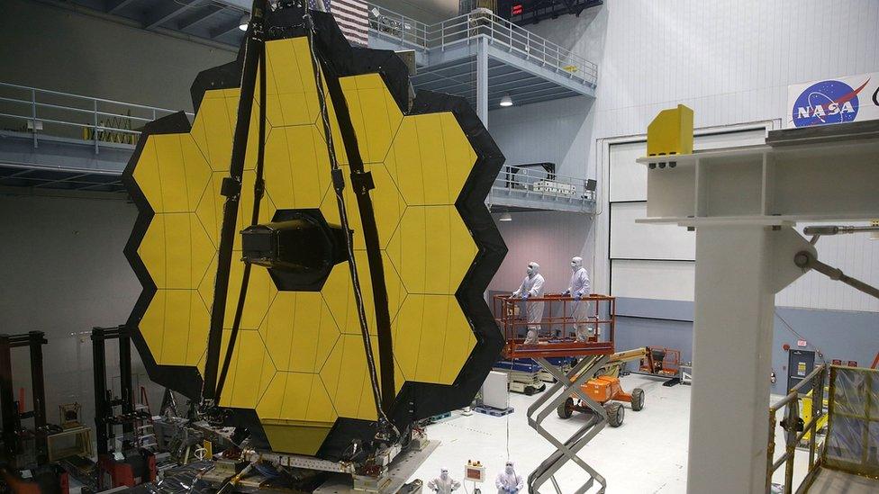 A large assemblage of gold hexagons, in a clean room workshop with suited workers looking on