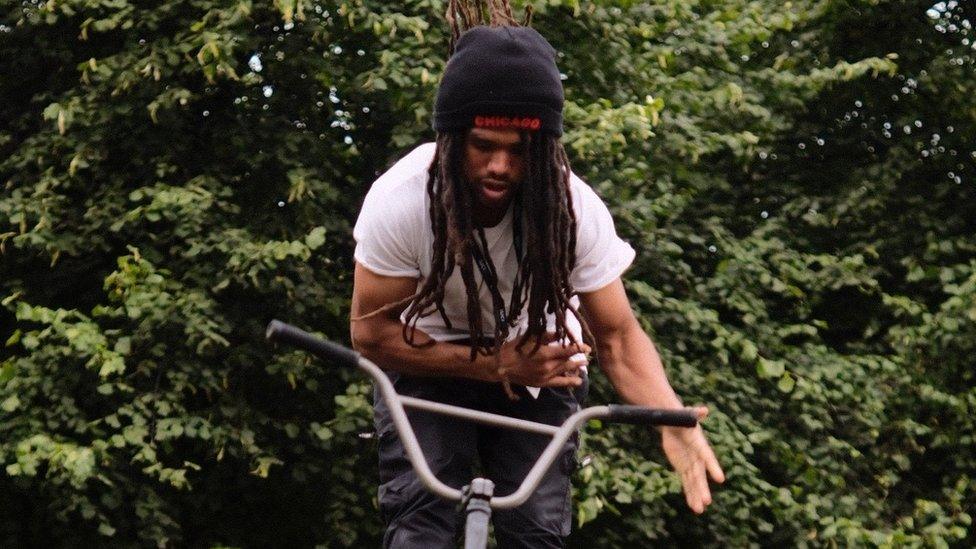 Andre Gillies, a black man in his early 20s, is pictured riding hands free on his BMX bike. Andre wears a black beanie hat with 'Chicago' stitched in red on the front. His hat covers his long dread locks down to his middle. He also wears a plain white t-shirt and black jeans. He is looking down at his handle bars and has a focussed expression with his mouth slightly open. He is photographed outside in front of a green hedge which fills the backdrop