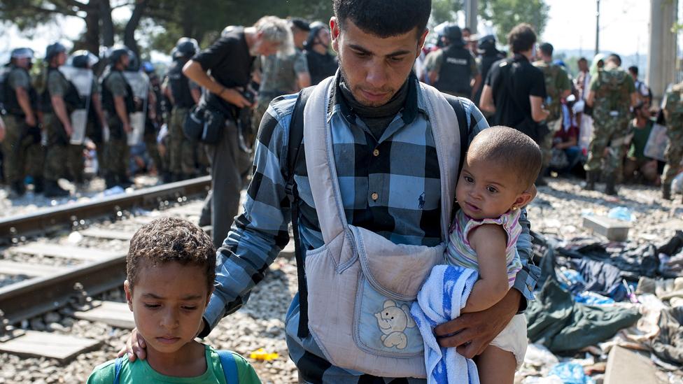 A man walks with children as police officers stand at the border line between Greece and Macedonia near the town of Gevgelija on August 26, 2015.