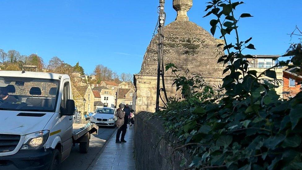 A pedestrian on the pavement on the tight medieval bridge in the town, with cars going past