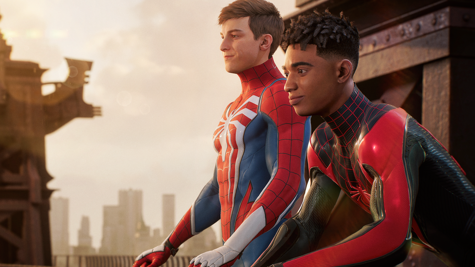 A screenshot from Spider-Man 2 showing protagonists Peter Parker and Miles Morales sitting on a balcony. They're both wearing their figure-hugging Spidey Suit costumes, but neither is wearing a mask. Peter's suit is mostly red, with thin black lines suggesting a spider's web. It also has blue panels on the biceps, triceps and flanks of his torso. A large, white, spider decal is printed on his chest, its legs extending to the edges of the suit. Peter is white, with fairly short, neatly cropped hair. Mile's suit has a similar design but is mainly black, with red panelling, web lines and spider decal. He's a young black man with short, afro-style hair style in locs. Both look out over New York contemplatively.