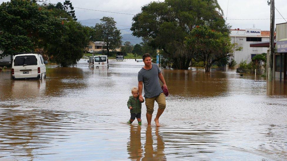 A man and a young boy walk through shallow floodwaters.