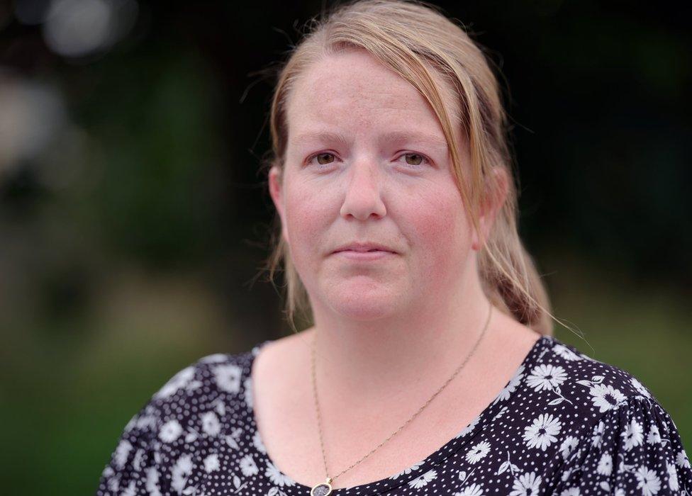 Jodie is outside in front of a blurred out tree. She is wearing a black and white patterned dress and a gold necklace. She has a concerned expression.