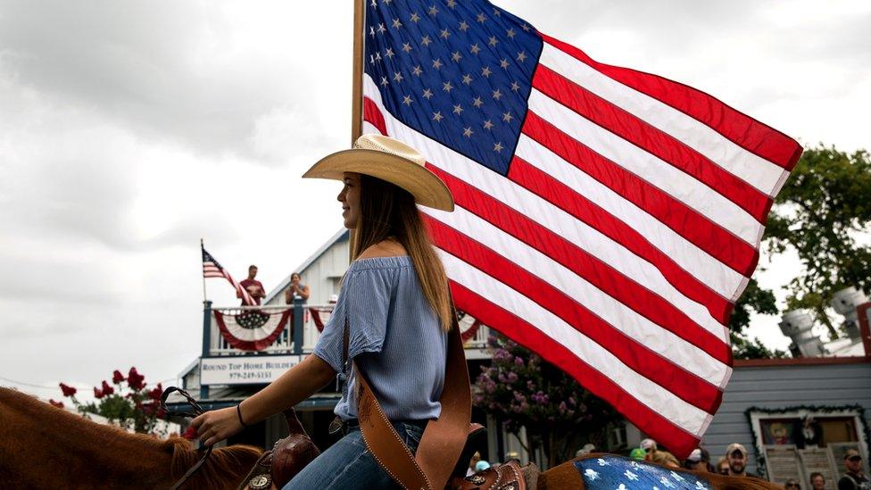 Girl pictured in profile on a horse, wearing a cowboy hat and carrying an American flag