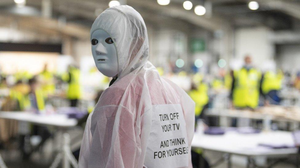 A person dressed in a white hazmat suit and a white mask, they have a sign attached to their back saying "Turn off your TV, think for yourself."