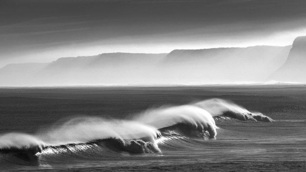 black and whiite photograph of waves rolling along the surface of the sea with cliffs shrouded in mist in the background