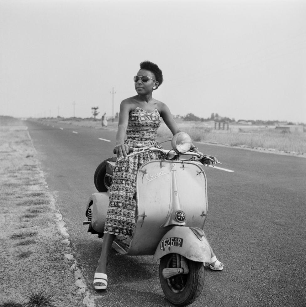 A woman sitting on a moped by the roadside poses for the camera.