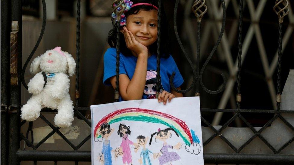 A little girl in Indonesia holds up a drawing of her family while she's stood behind a gate