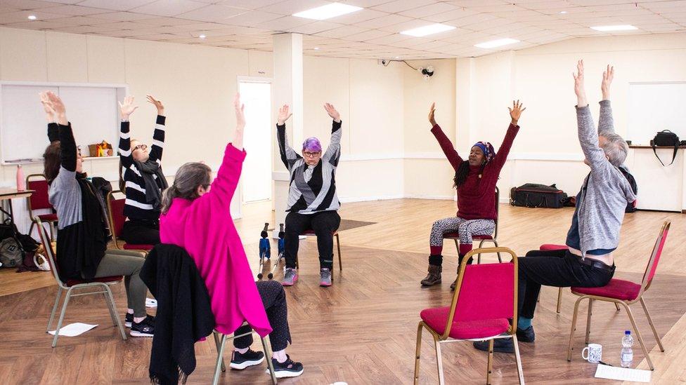 People dancing in their chairs, arranged in a circle, in a hall